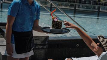 A person holding a tray with a pineapple drink and a small umbrella, standing by a pool.