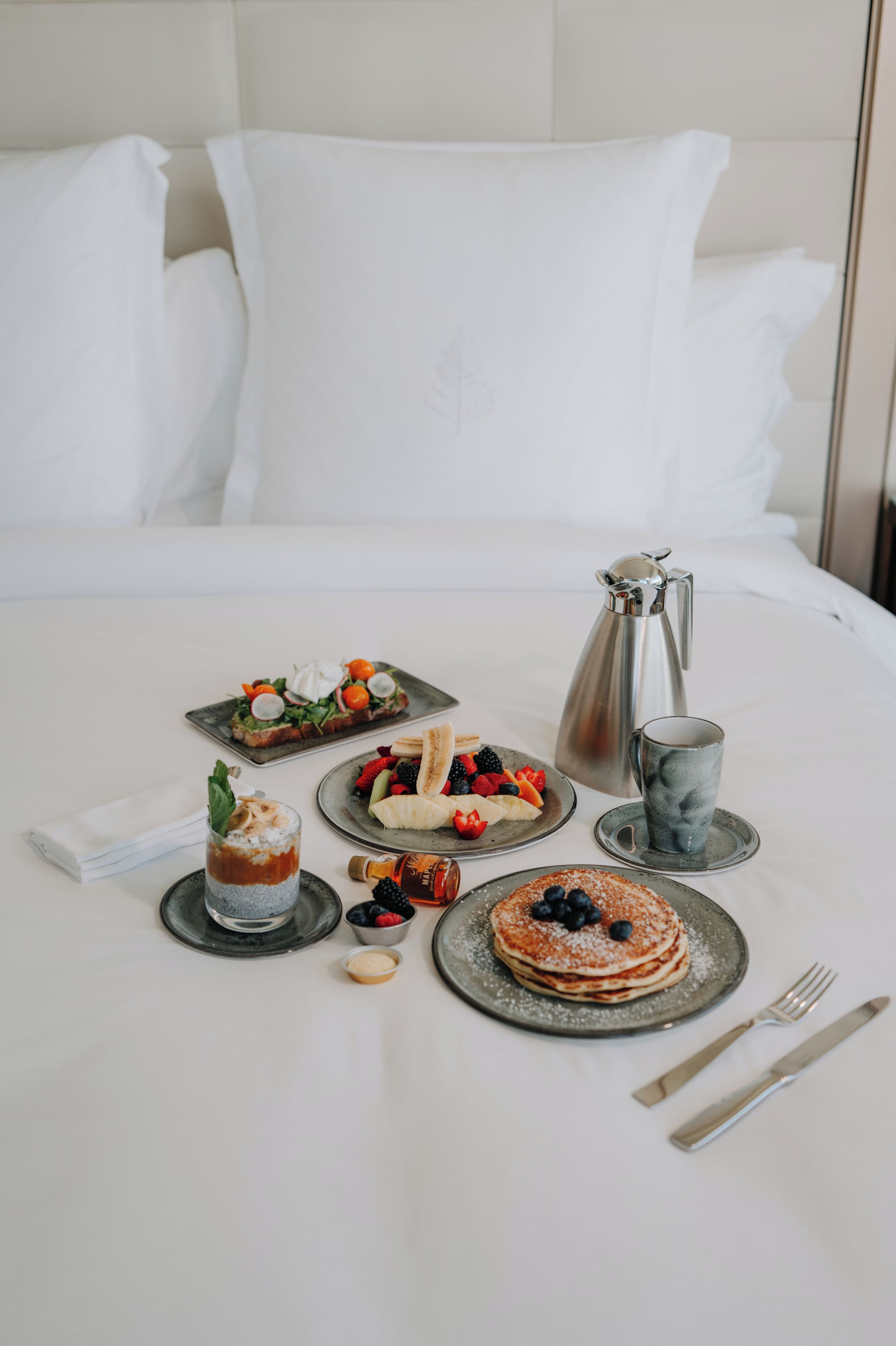 A neatly arranged breakfast buffet on a hotel bed with a variety of dishes, a coffee pot, and dining utensils.
