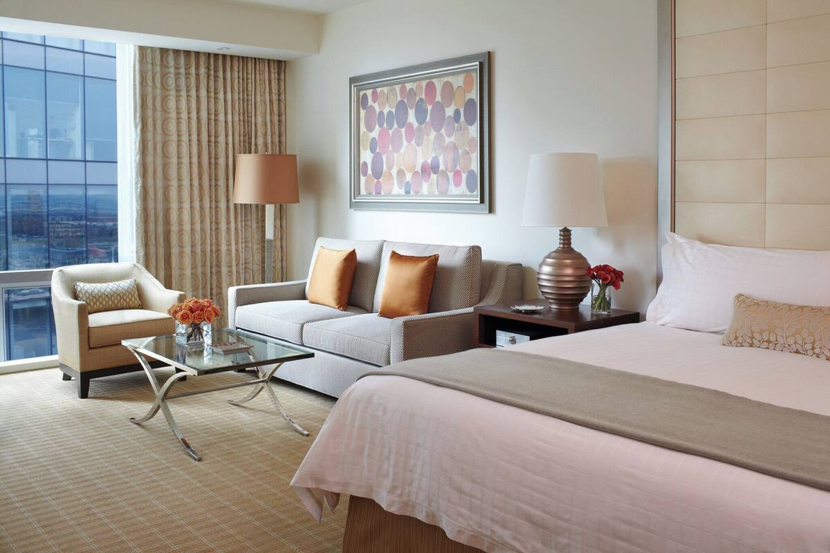 A hotel room with a bed, a sofa, a chair, a coffee table, and a large window offering a city view.