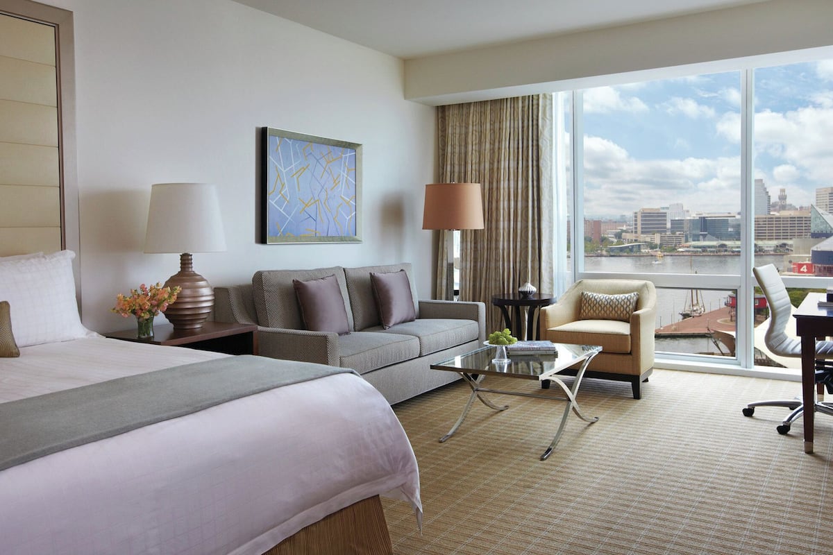A hotel room with a large bed, a sofa, a chair, a small table, and a view of a cityscape through large windows.