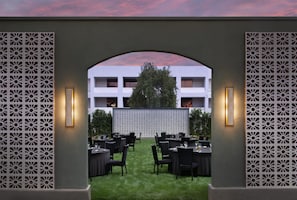 Uma área de refeições ao ar livre com mesas e cadeiras, um muro decorativo e um edifício ao fundo.