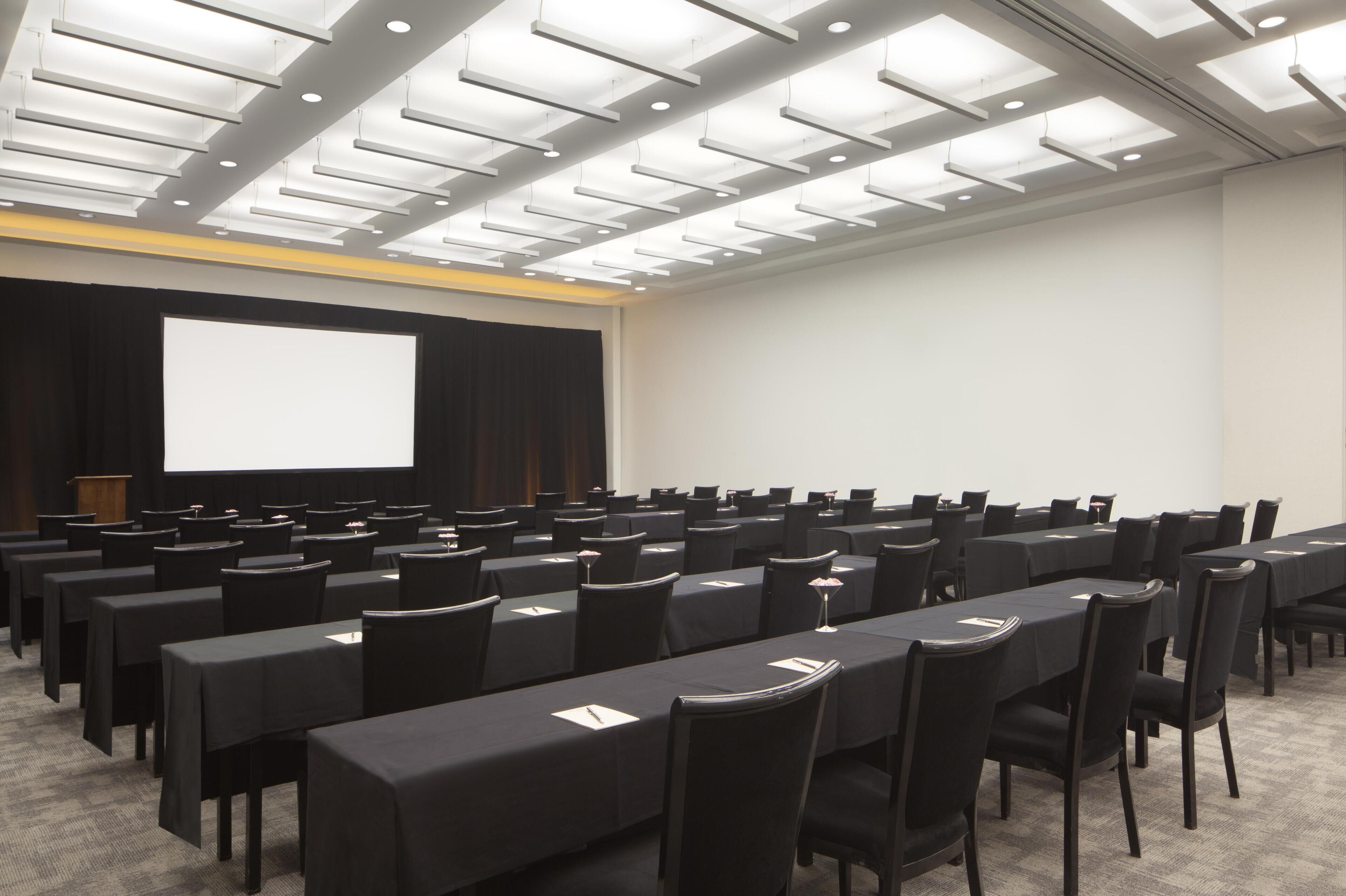 Un salón de conferencias con mesas, sillas, una pantalla de proyector y un atril.