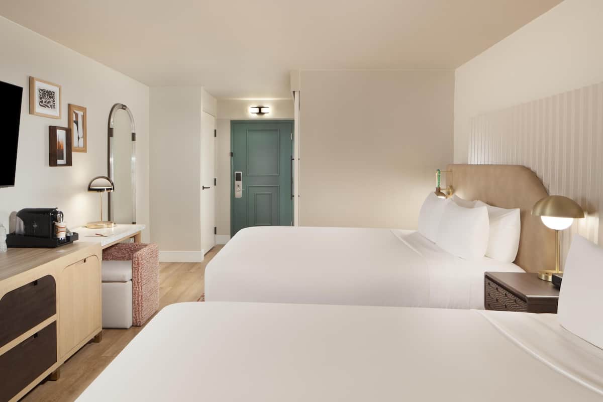 A hotel room with two beds, a desk with a coffee maker, a TV, and a door leading to another room.
