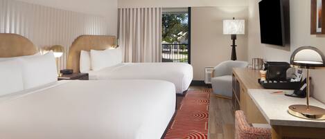 A hotel room with two beds, a desk, a chair, a TV, and a view of the outdoors.