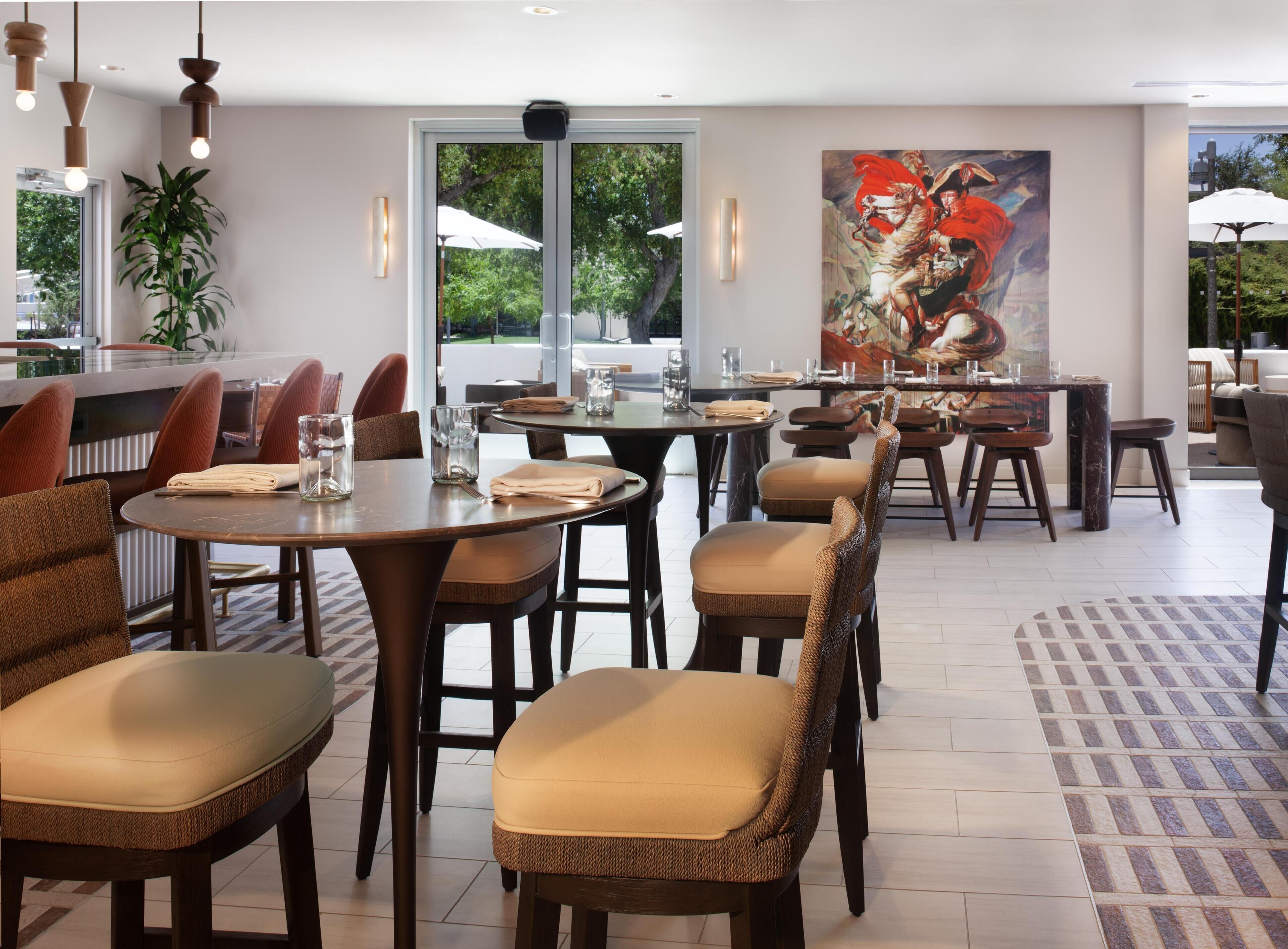 A restaurant with round tables and cushioned chairs, a large painting on the wall, and a view of greenery outside.