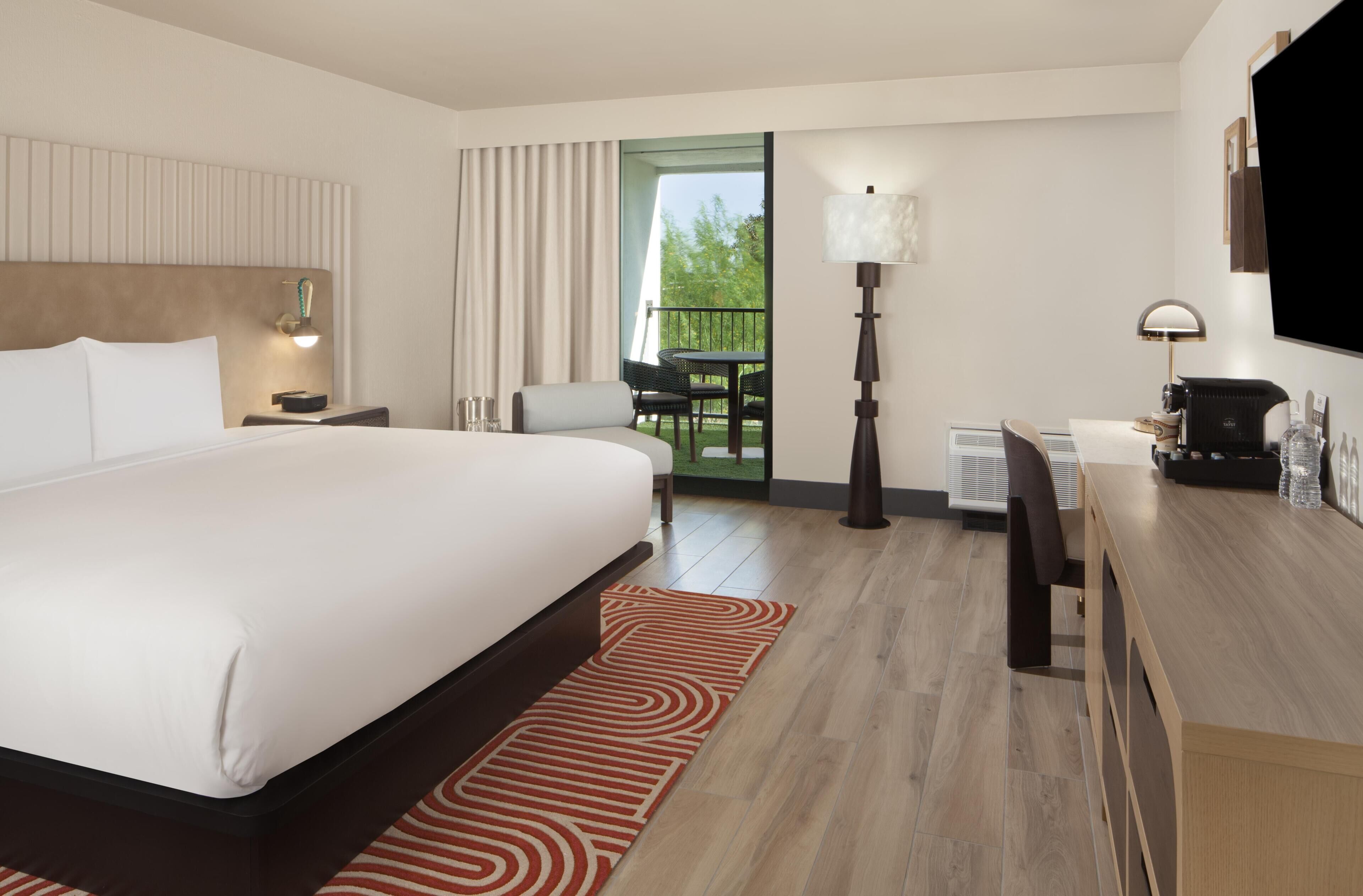 A hotel room with a large bed, a desk, a chair, a TV, and a balcony with a view.