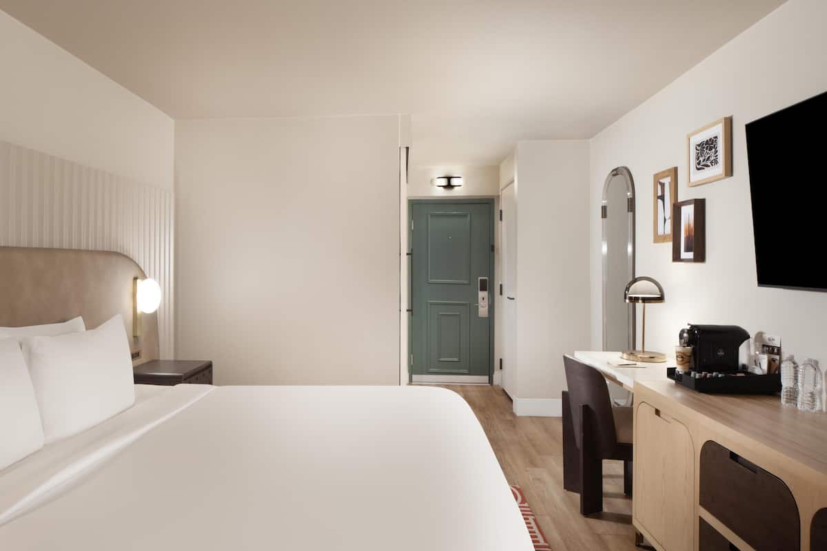 A hotel room with a bed, bedside table, desk, chair, and a door leading to another room.