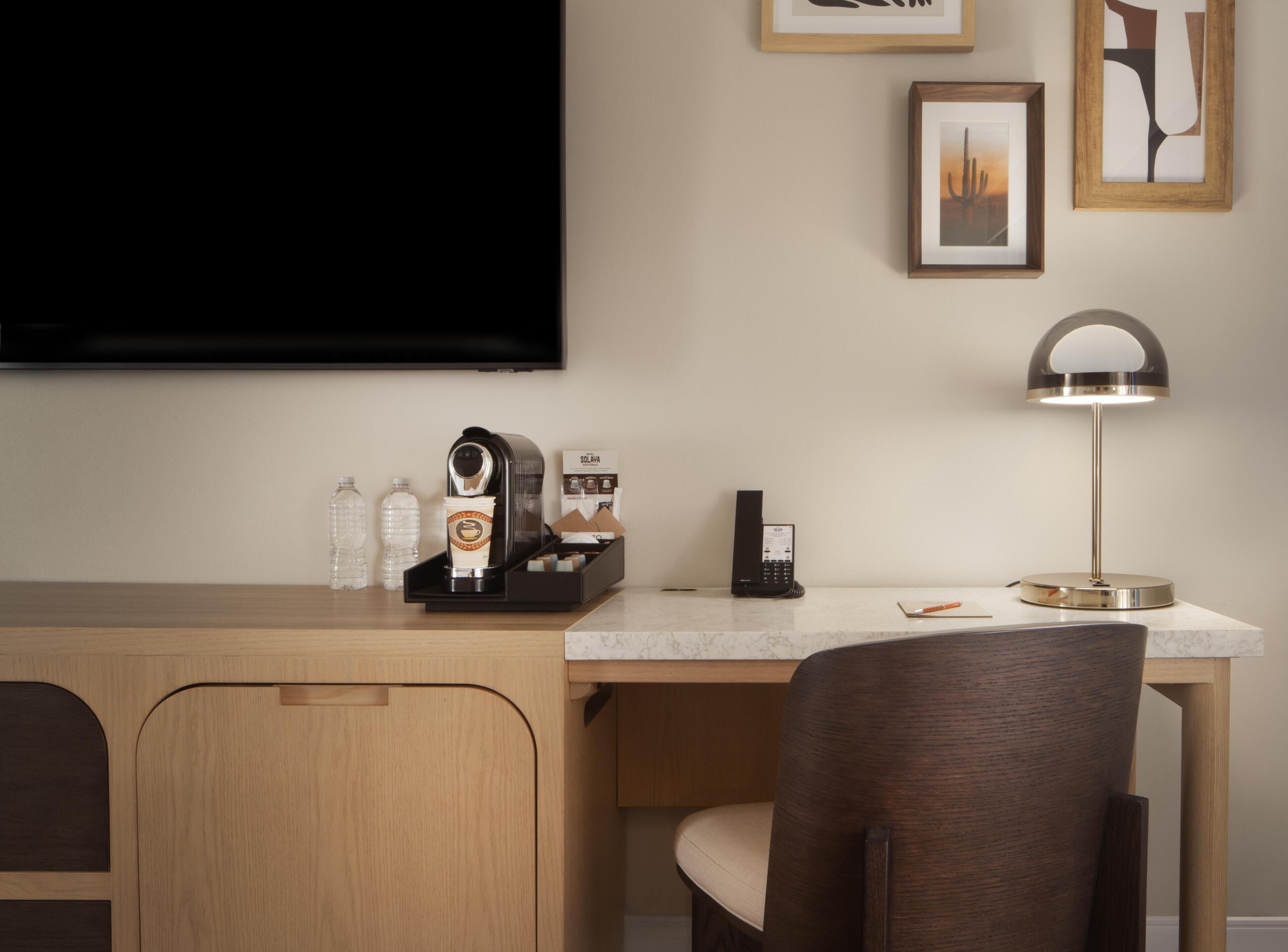 Hotelkamer met een houten bureau, een koffiezetapparaat, telefoon, lamp en lijstjes met kunstwerken aan de muur.