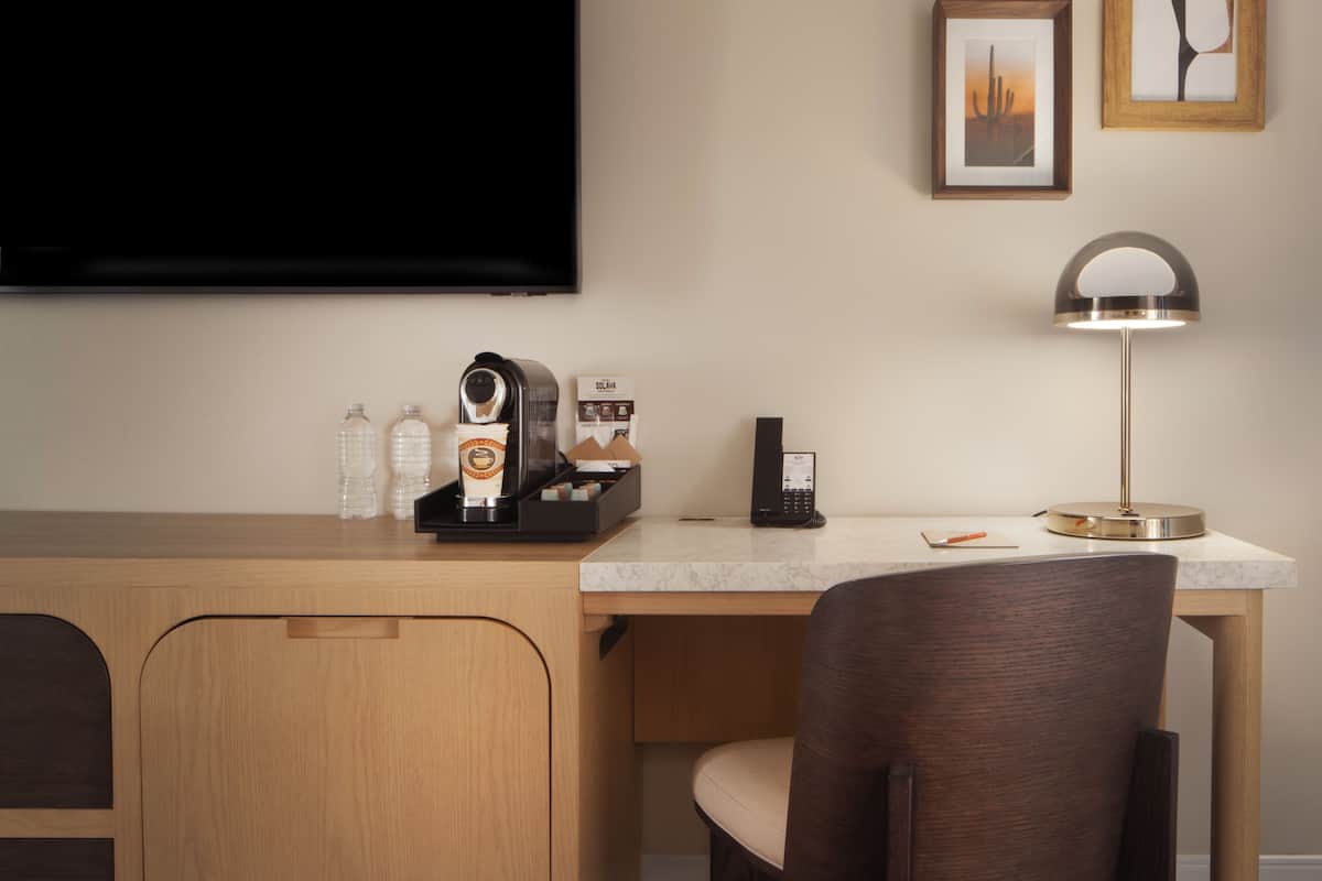 A hotel room with a wooden desk, a coffee machine, a phone, a lamp, and a TV mounted on the wall.