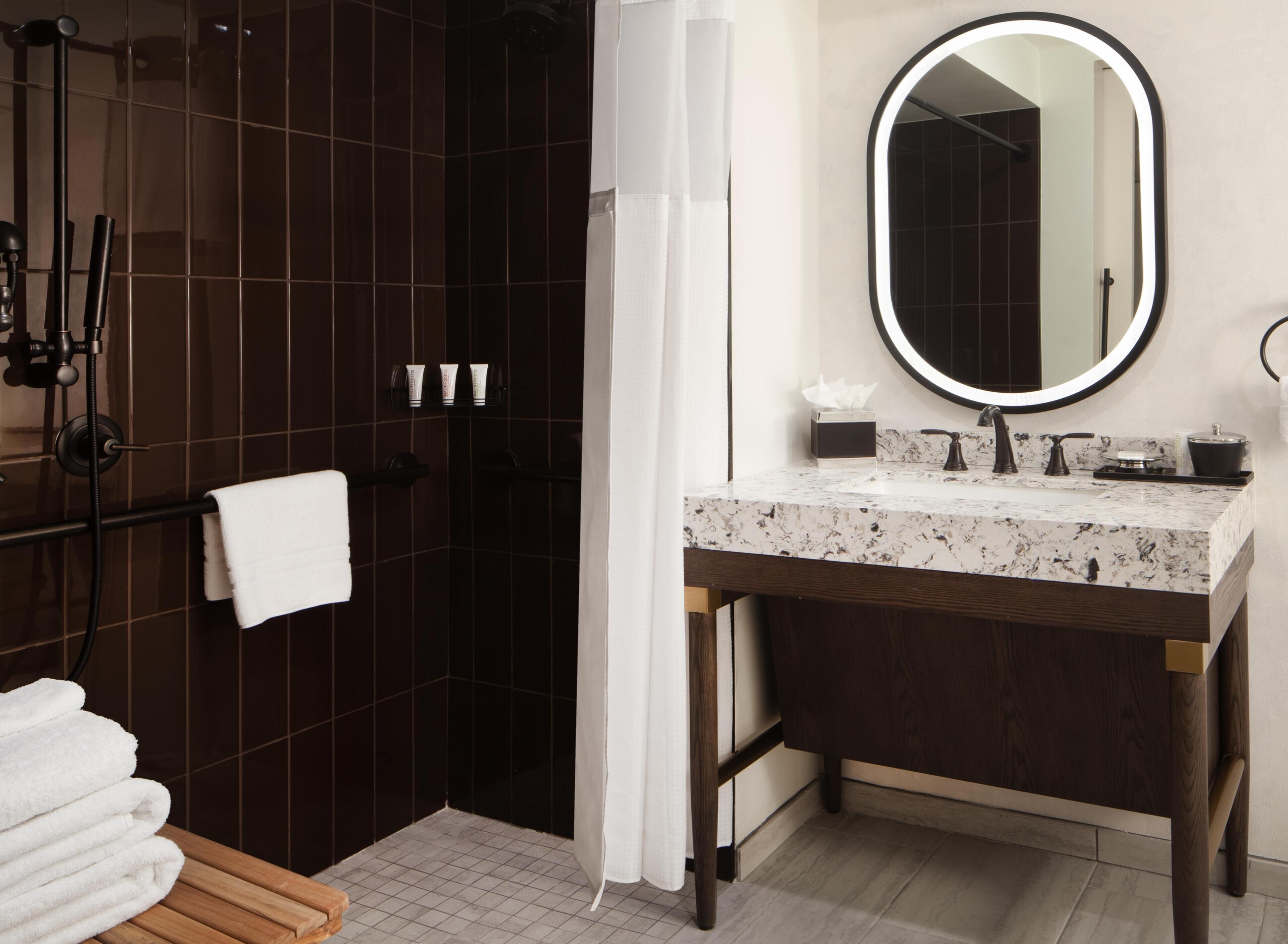 A bathroom with a dark tiled shower, a white towel on a rack, a wooden bench, a marble sink with a mirror, and toiletries on the counter.