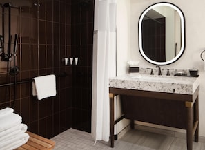 A bathroom with a dark tiled shower, a white towel on a rack, a wooden bench, a marble sink with a mirror, and toiletries on the counter.