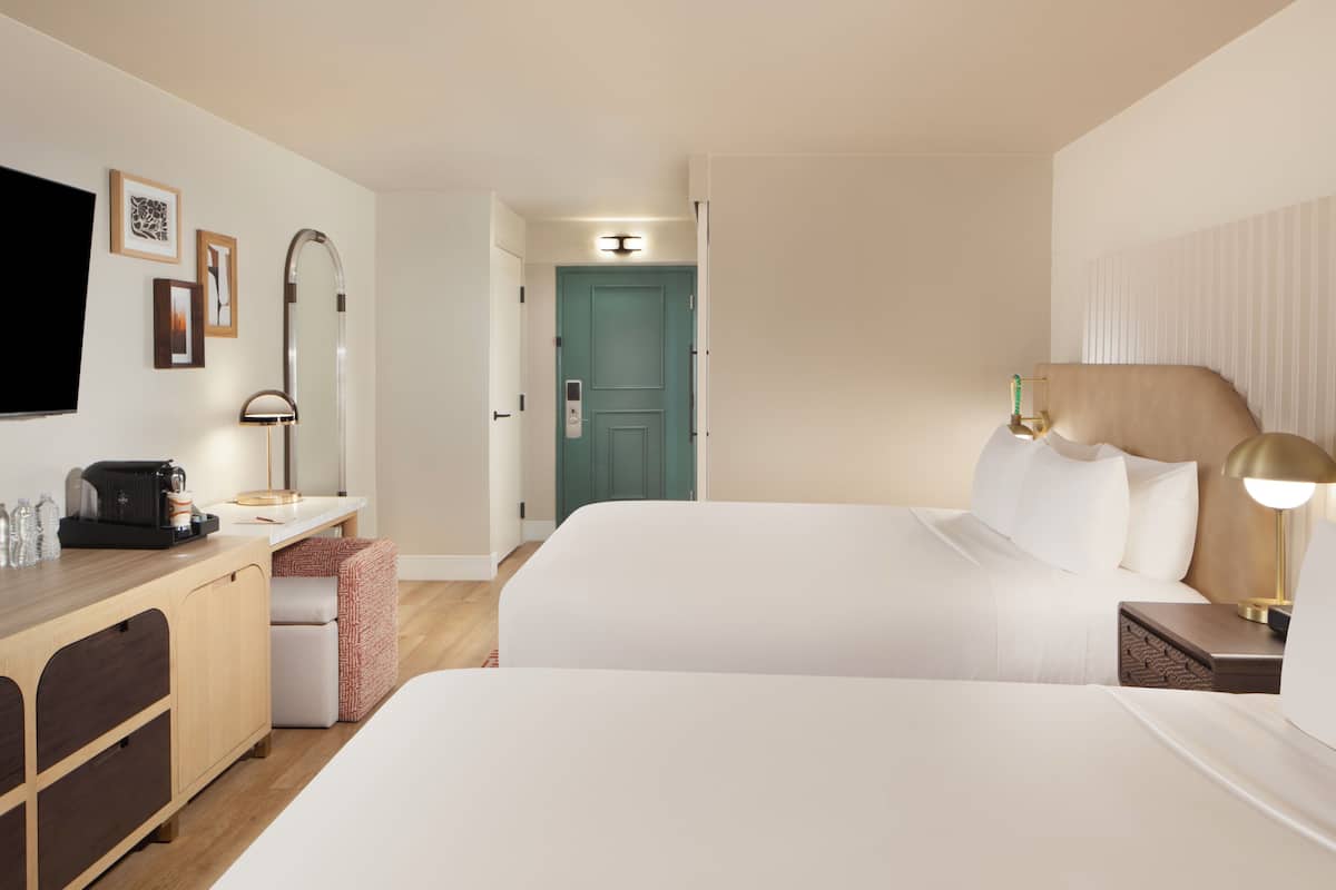 A hotel room with two beds, a desk with a coffee maker, a TV, and a door leading to another room.