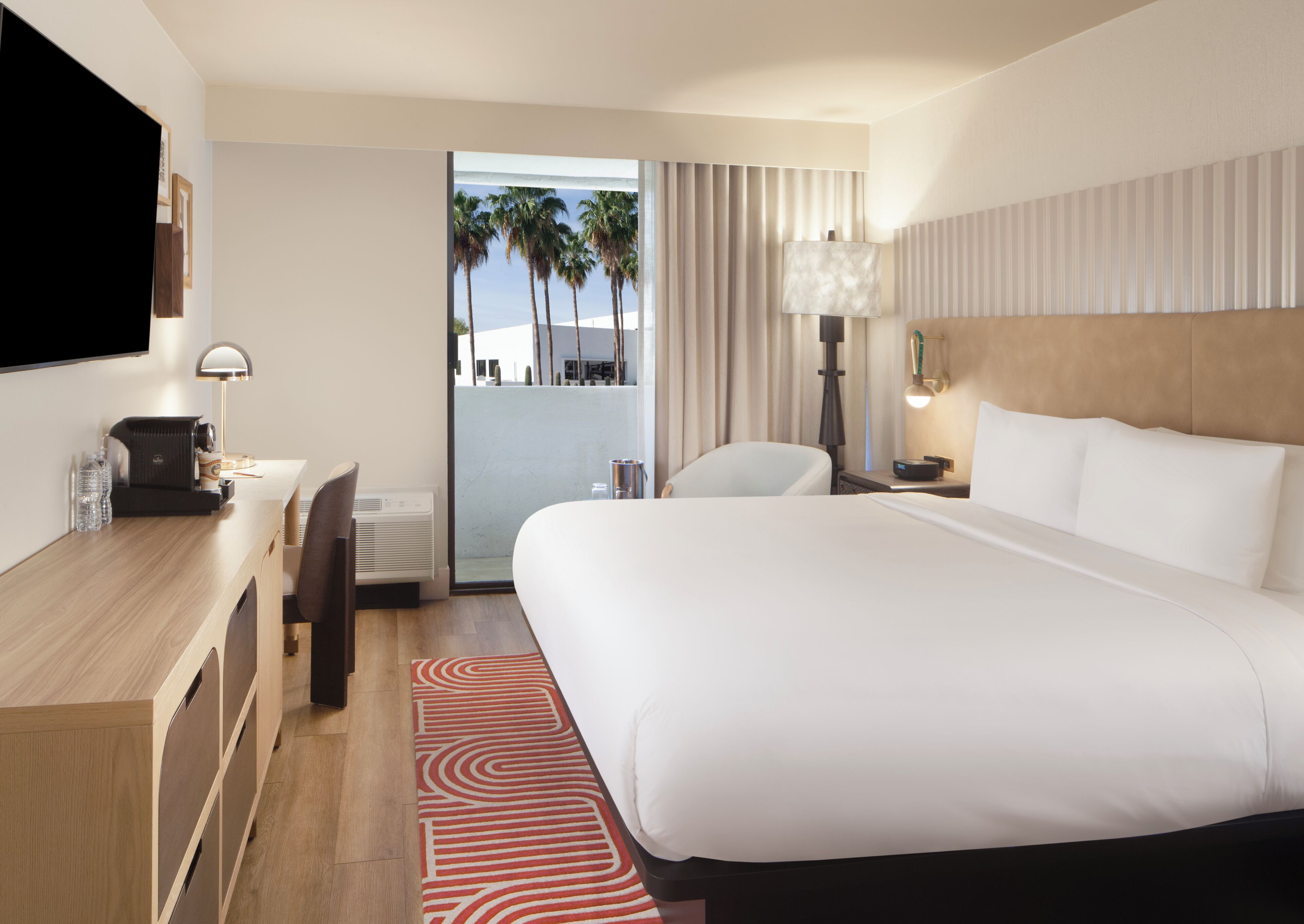 A hotel room with a bed, desk, chair, TV, lamp, and a view of palm trees.