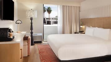 Habitación de hotel con una cama grande, un escritorio con cafetera, televisión y vista a palmeras.