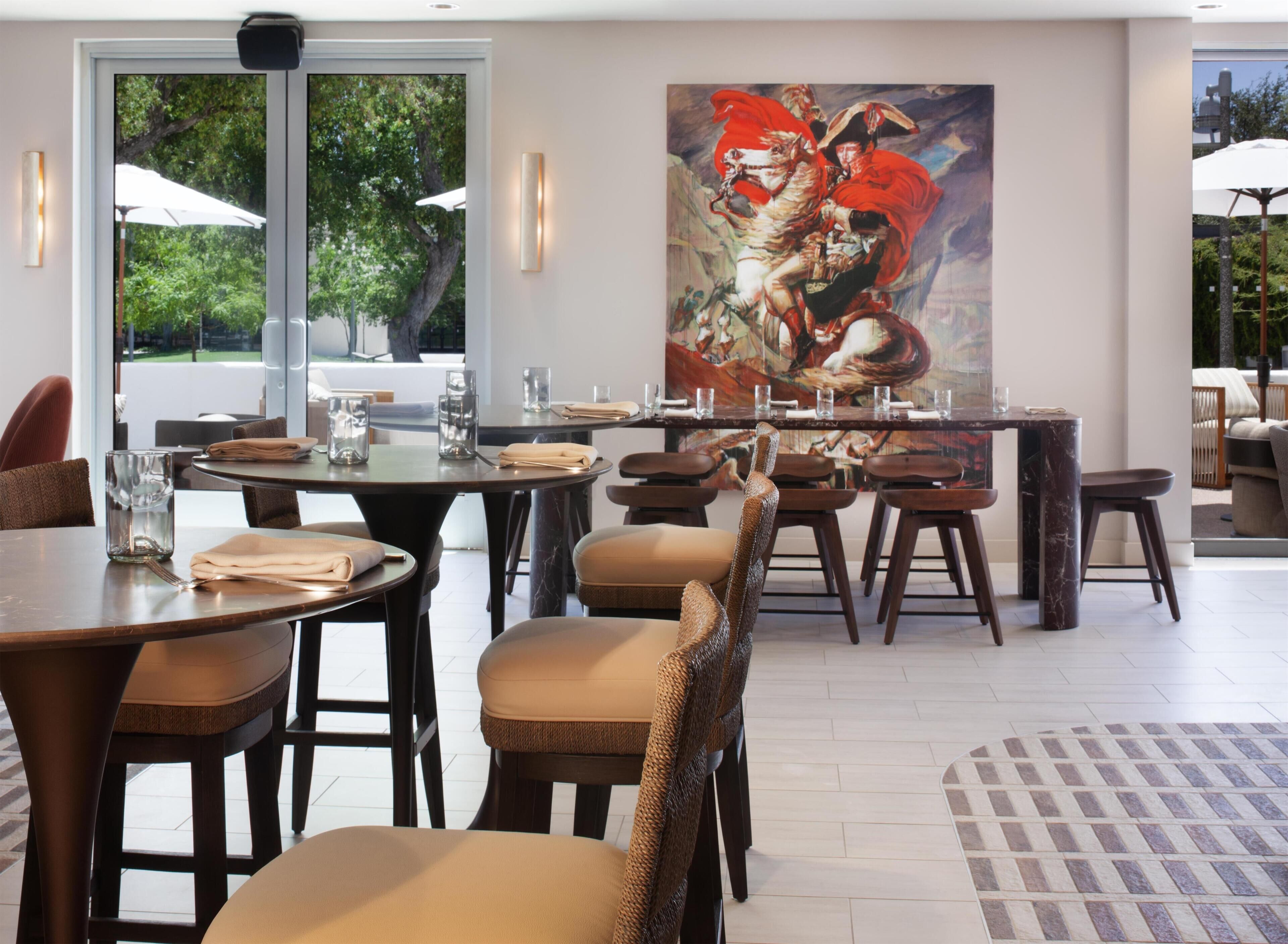 A restaurant with round tables, cushioned chairs, and a large painting on the wall.