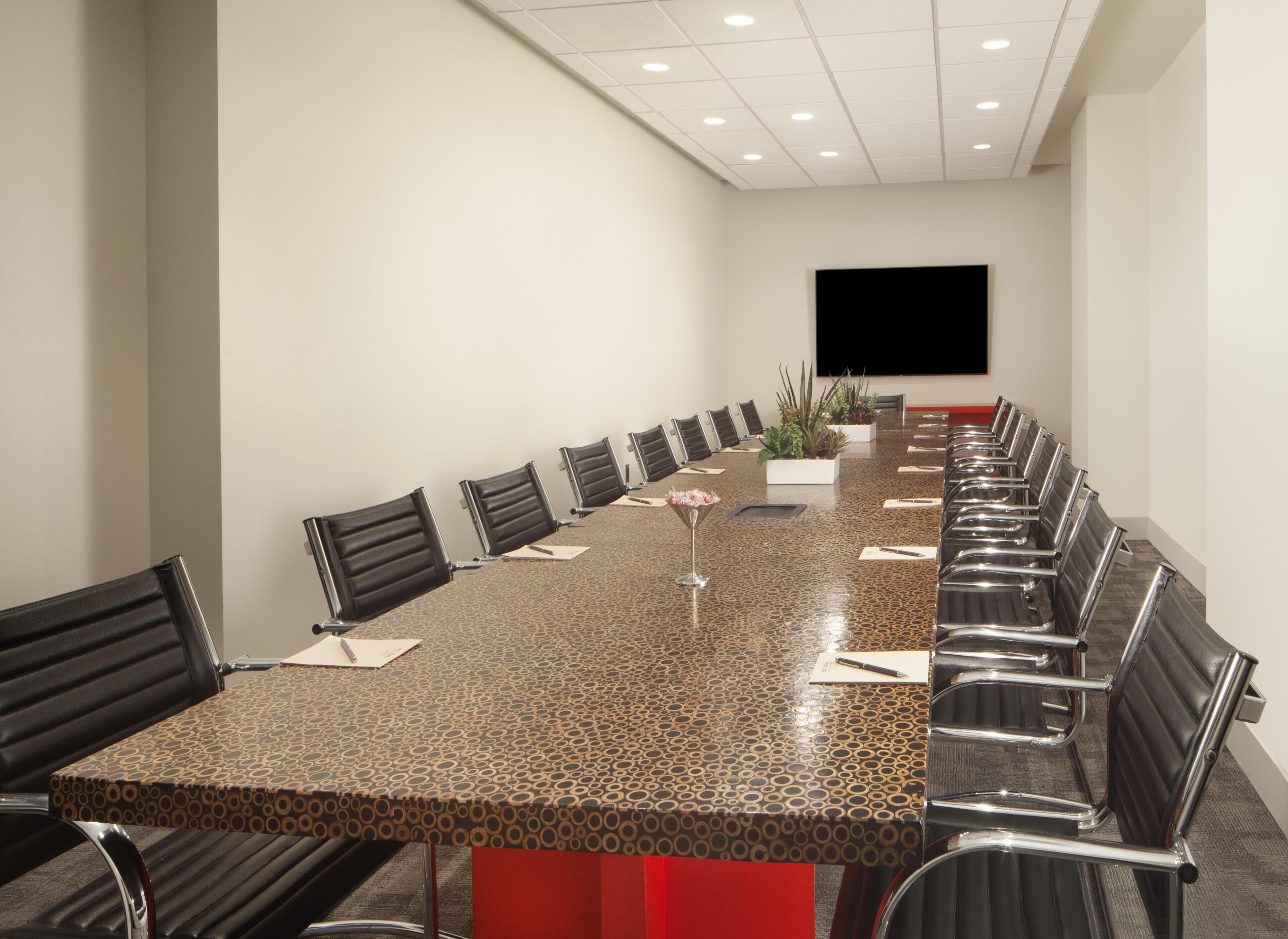 A conference room with a long table, chairs, a plant, and a screen.