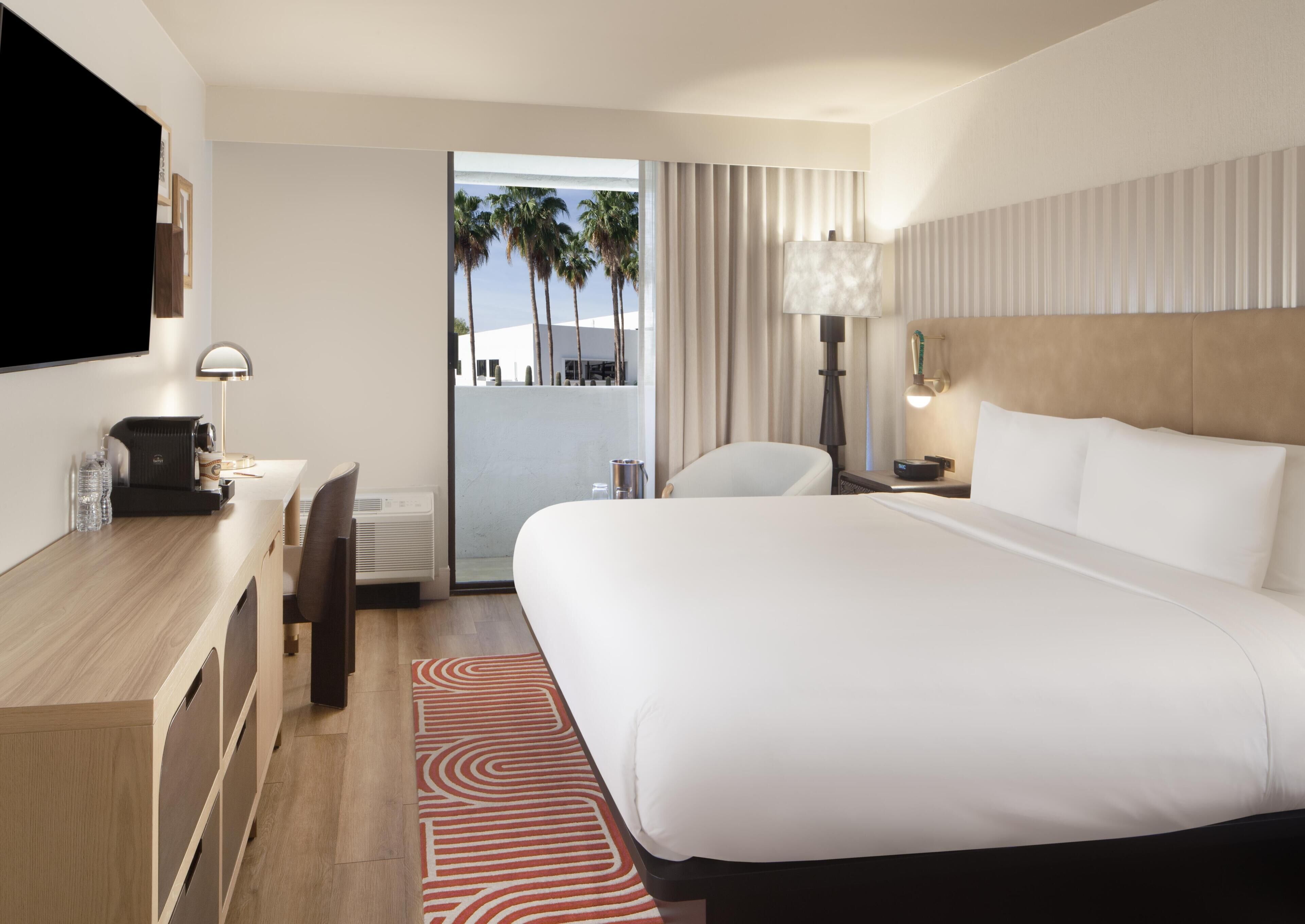 A hotel room with a bed, desk, chair, lamp, and a view of palm trees.