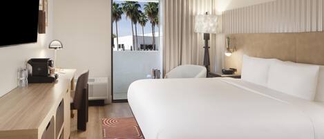 A hotel room with a bed, desk, chair, lamp, and a view of palm trees.