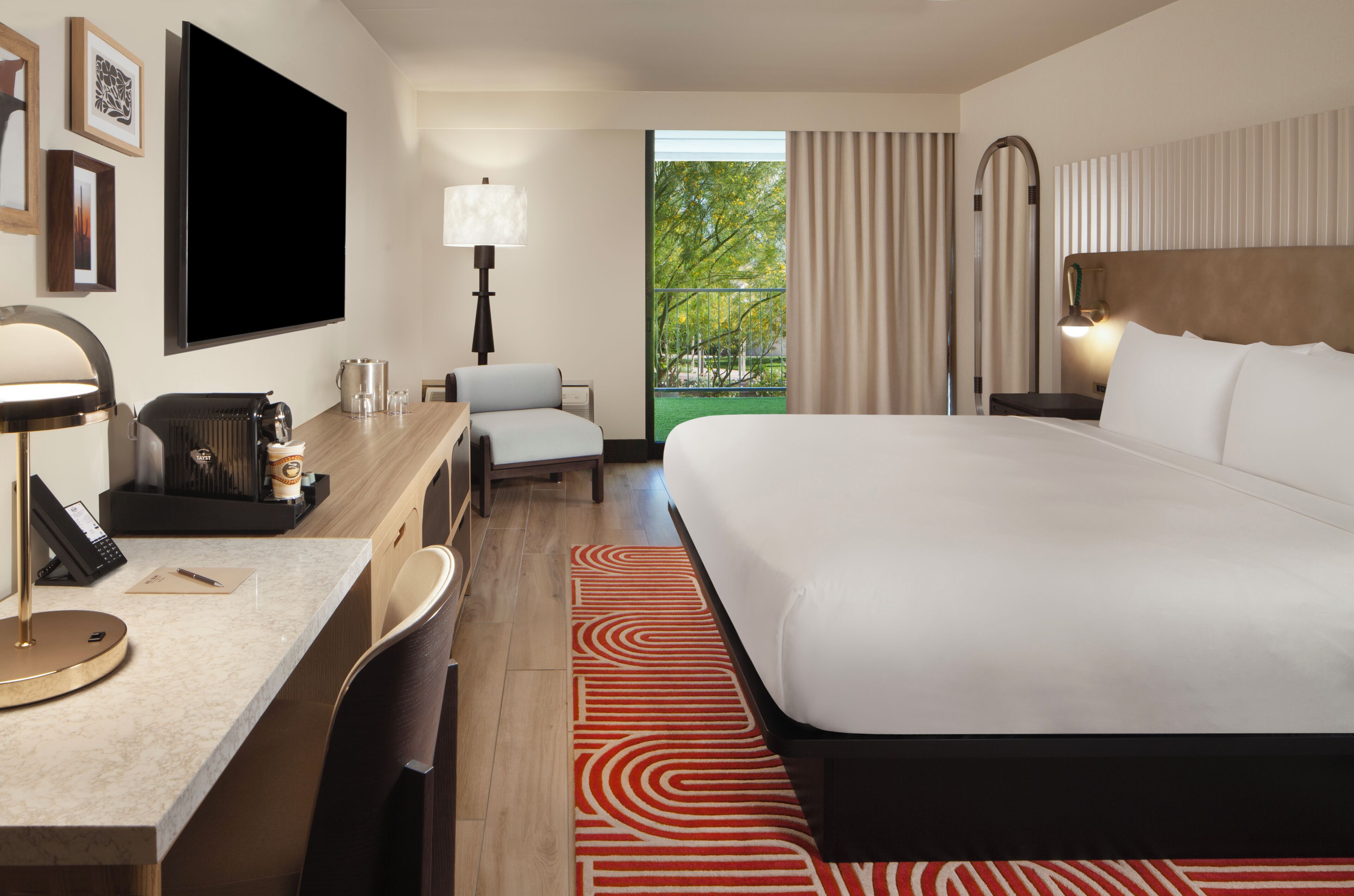 A hotel room with a large bed, a desk with a lamp, a chair, a TV, and a view of greenery outside.