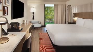 A hotel room with a large bed, a desk with a lamp, a chair, a TV, and a view of greenery outside.