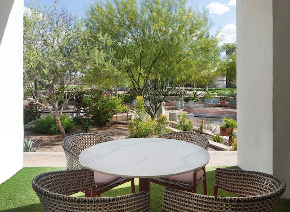 A patio with a round table and wicker chairs, surrounded by greenery and a view of a pool and buildings.