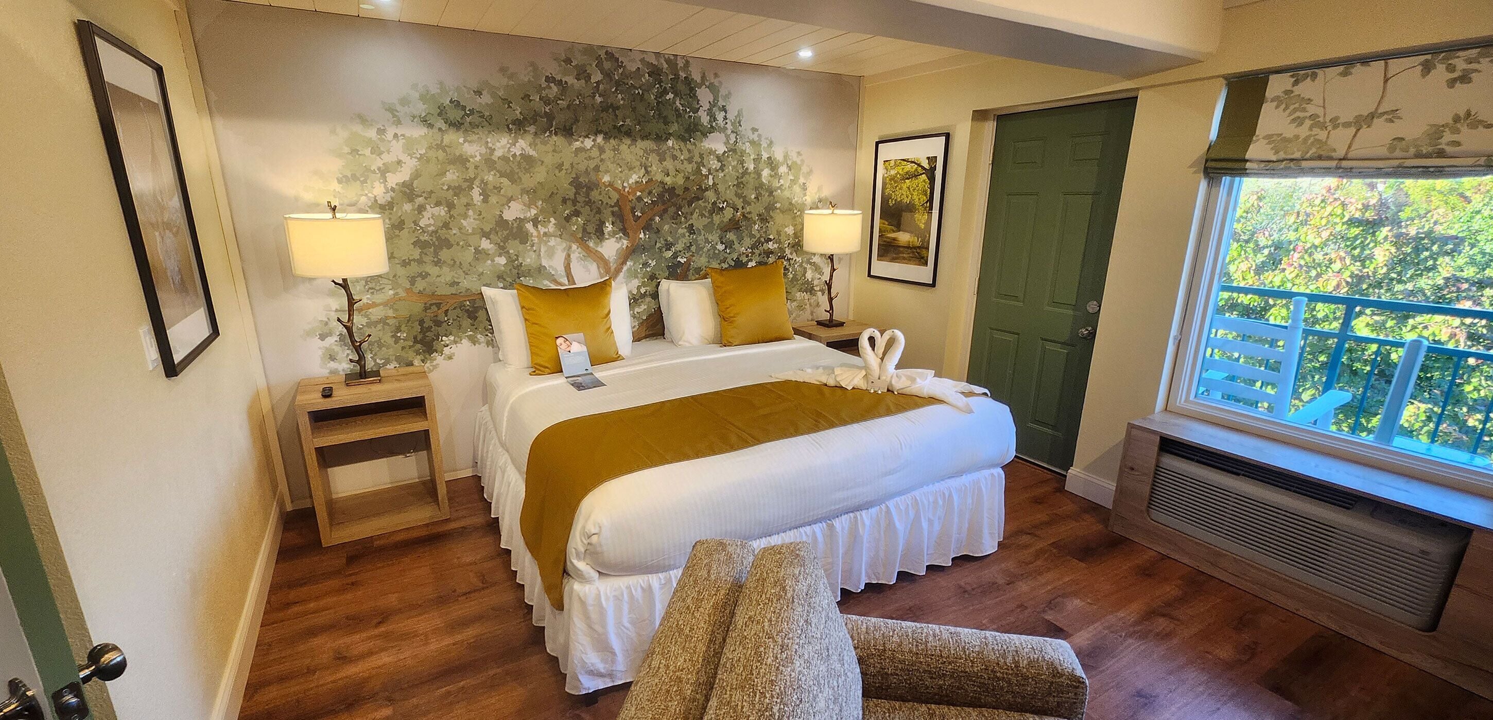 A bedroom with a large bed, a nightstand, a lamp, a chair, a green door, and a window with a view of trees.
