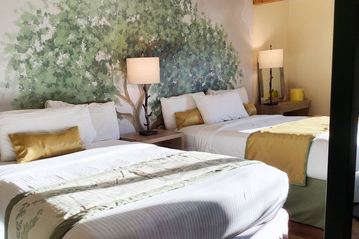A hotel room with two beds, each with white bedding and gold accents. There are two bedside tables with lamps, and a large wall mural of foliage.