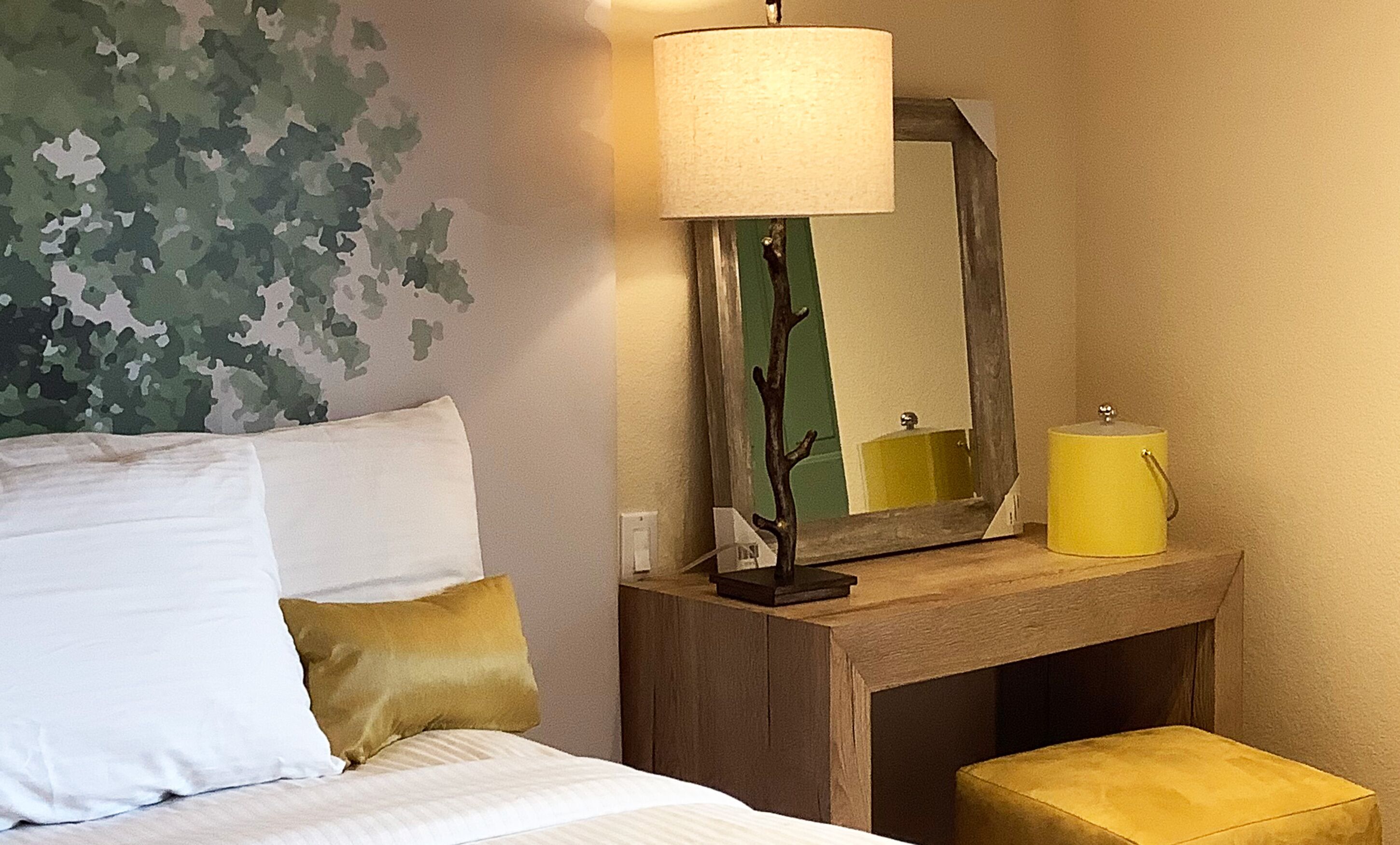 Un lit bien fait, agrémenté d’oreillers blancs et dorés, une lampe de chevet, un miroir et une petite table de chevet en bois sur laquelle se trouve un récipient jaune.