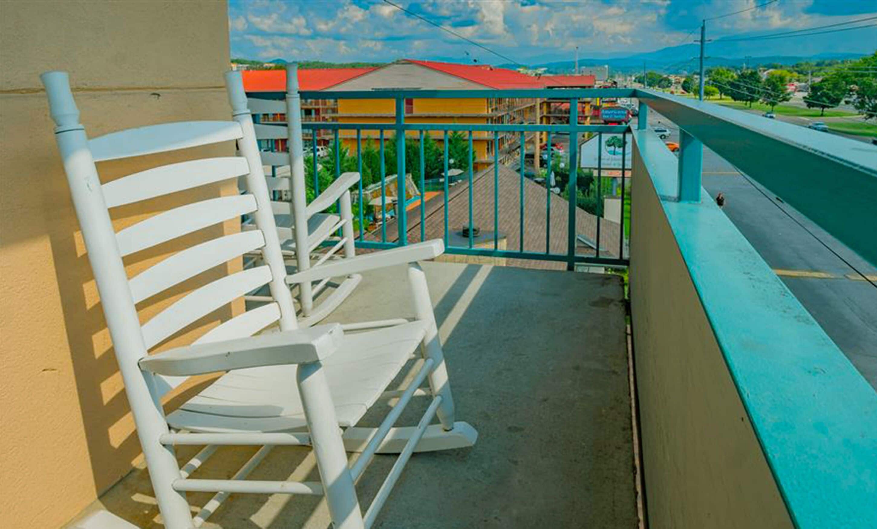 A balcony with white rocking chairs, a view of a street with buildings, and a clear sky.