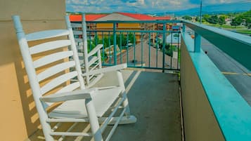 A balcony with white rocking chairs, a view of a street with buildings, and a clear sky.