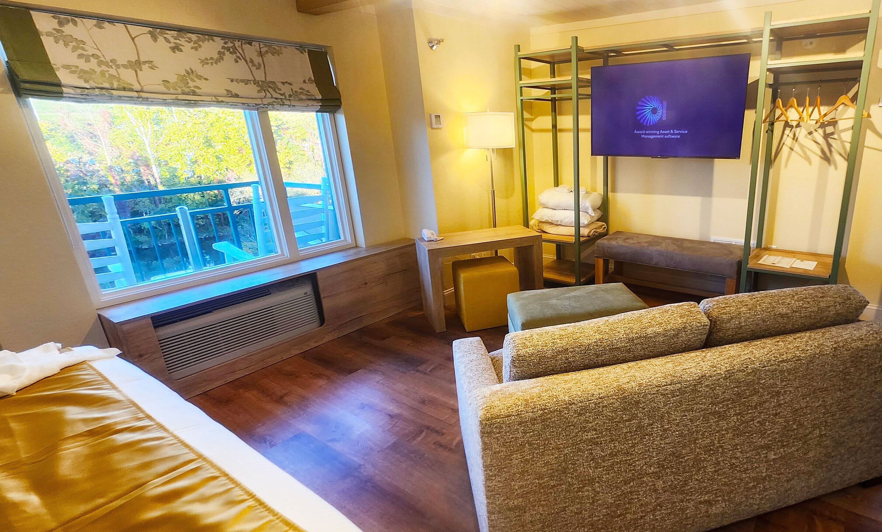 A hotel room with a bed, a sofa, a desk with a lamp, a television, and a window with a view of trees.