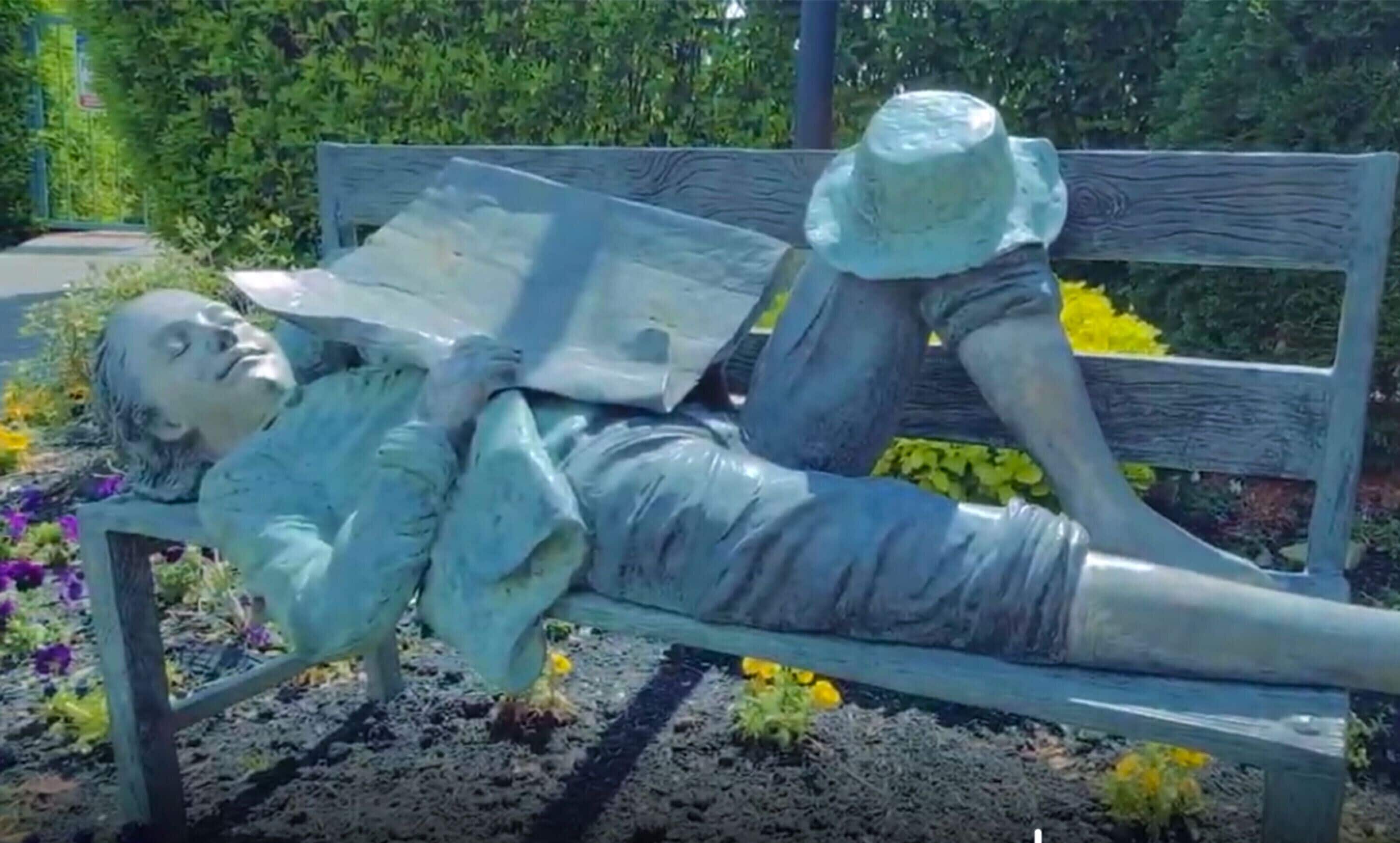 A statue of a person lying on a bench with a hat and a book.