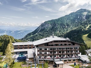 Un hôtel à l’architecture imposante, avec plusieurs étages, un espace extérieur aménagé pour s’asseoir et une vue sur un paysage montagneux.