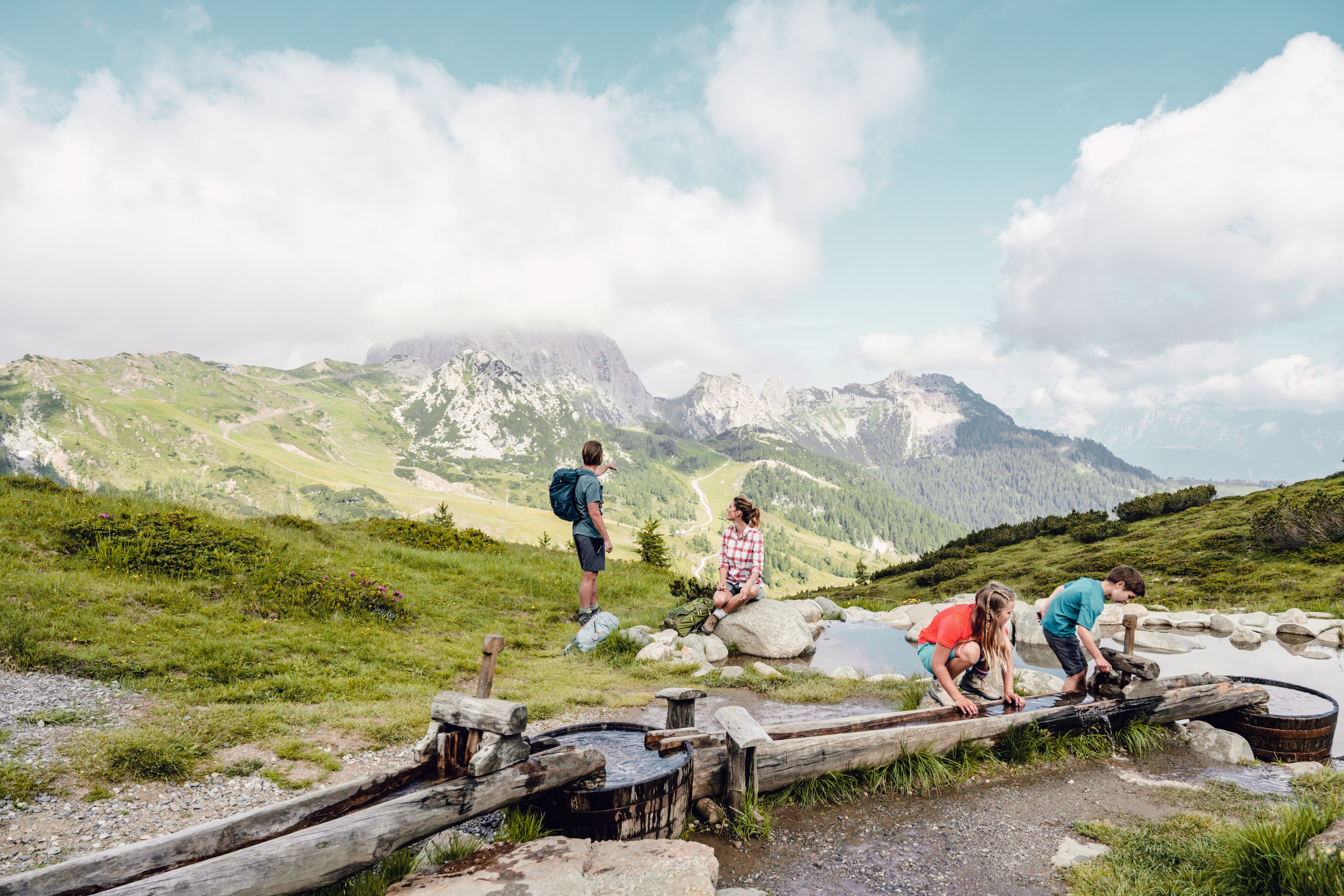 A group of people crossing a wooden bridge over a stream in a mountainous area with a clear sky.