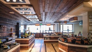 Ein modernes Restaurant mit Holzelementen, einer zentralen Anrichte mit Speisenpräsentationen und Blick auf die Küche im Hintergrund.