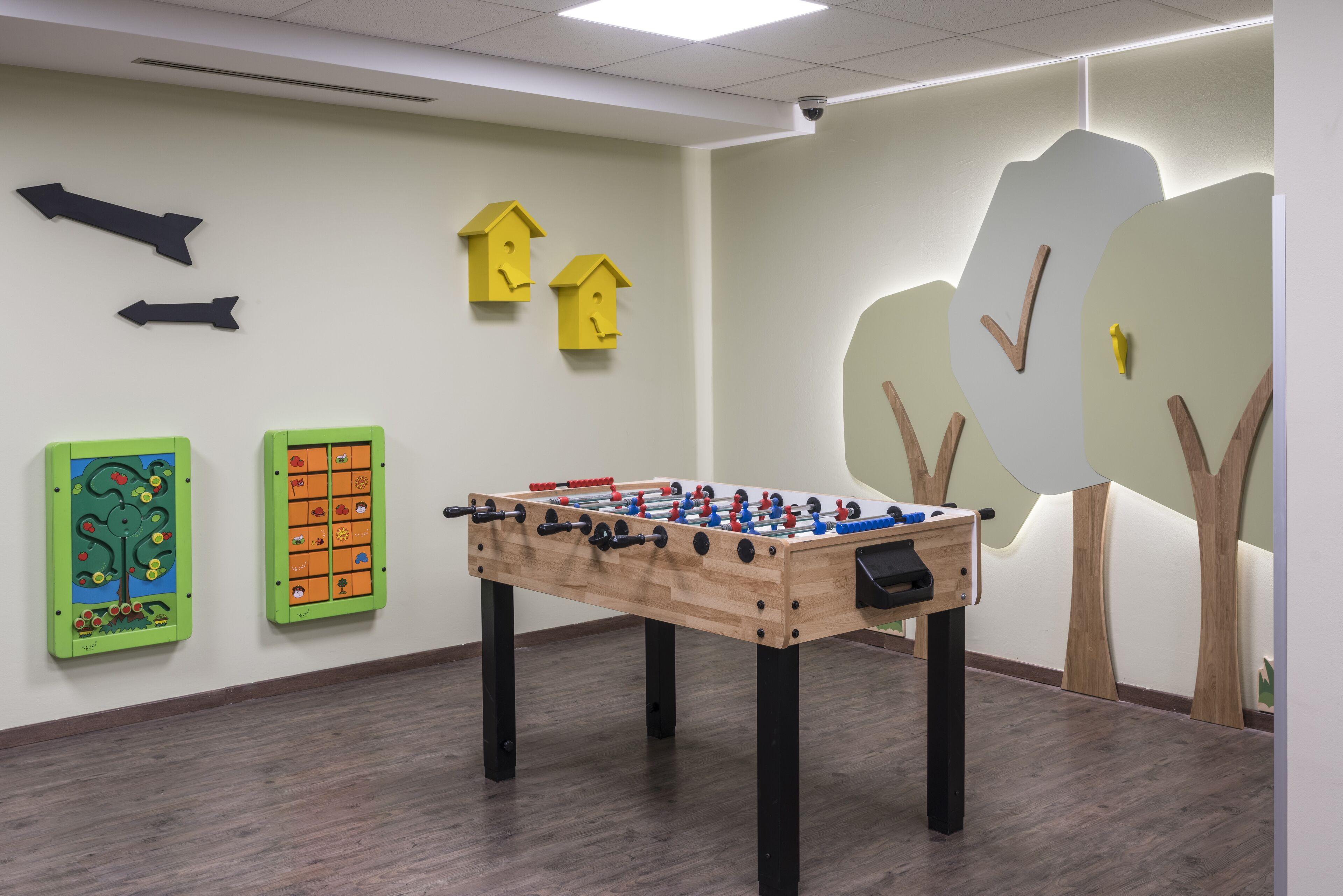 A room with a foosball table, two wall decorations resembling houses, and a wall-mounted camera.