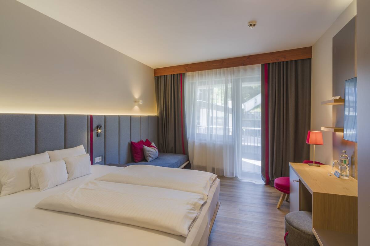 Habitación de hotel con dos camas, un escritorio, una silla y un gran ventanal con cortinas.