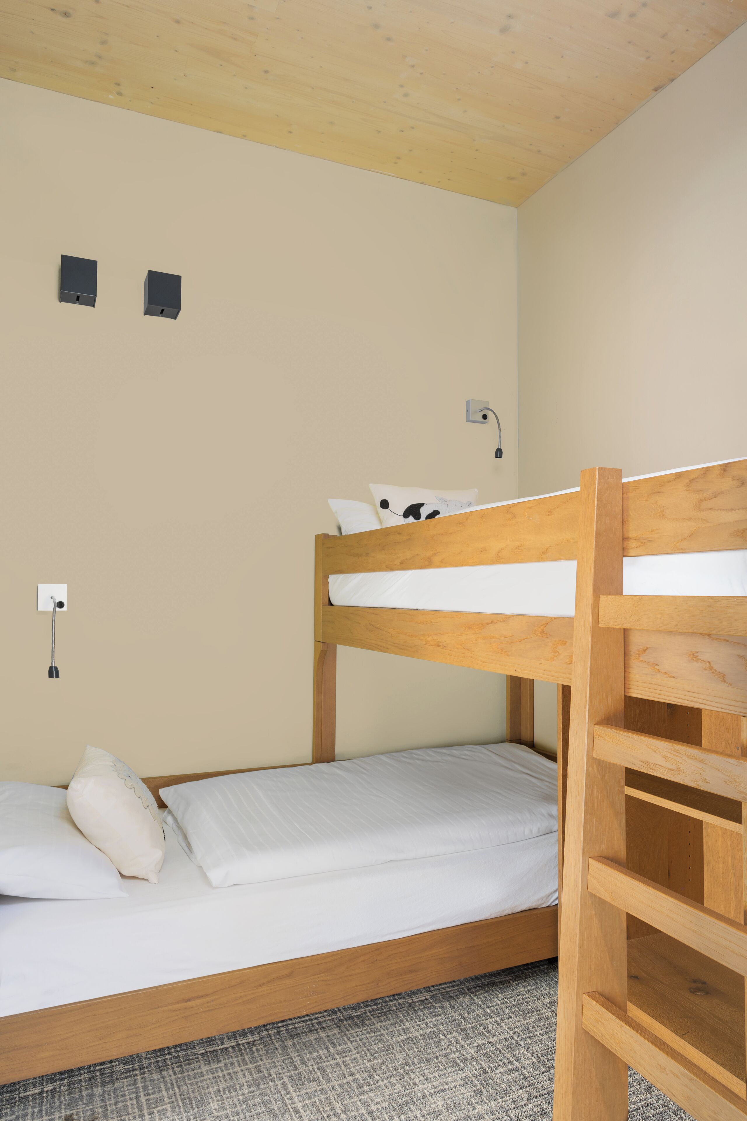 Una cama litera con estructura de madera, ropa de cama blanca y alfombra gris. Hay dos enchufes y un interruptor de luz en la pared.