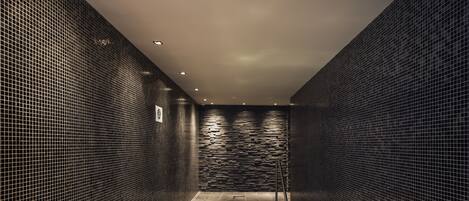Un moderno espacio de spa con una bañera de hidromasaje acristalada, barandillas metálicas y paredes de azulejos.