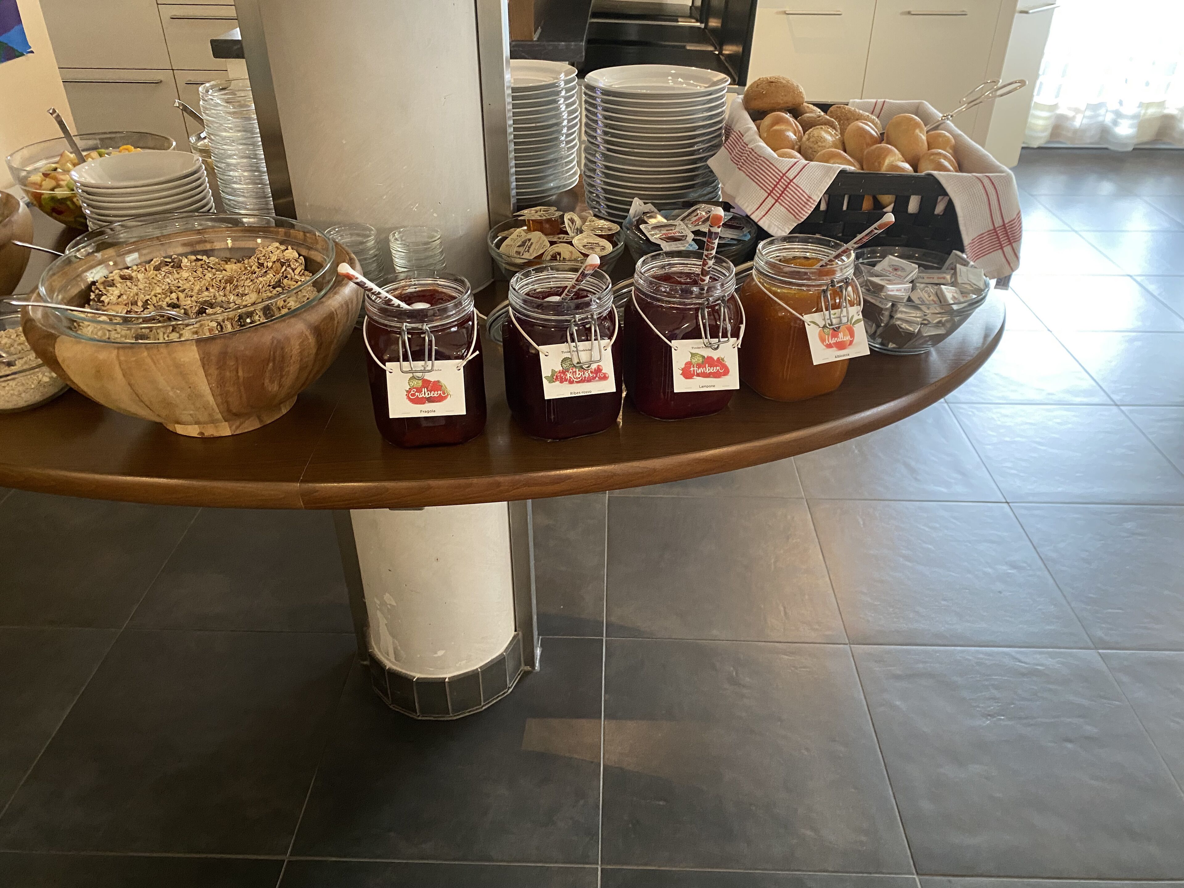 Una mesa de buffet con diversos alimentos, incluyendo frascos de mermelada, un tazón de granola y pilas de platos.
