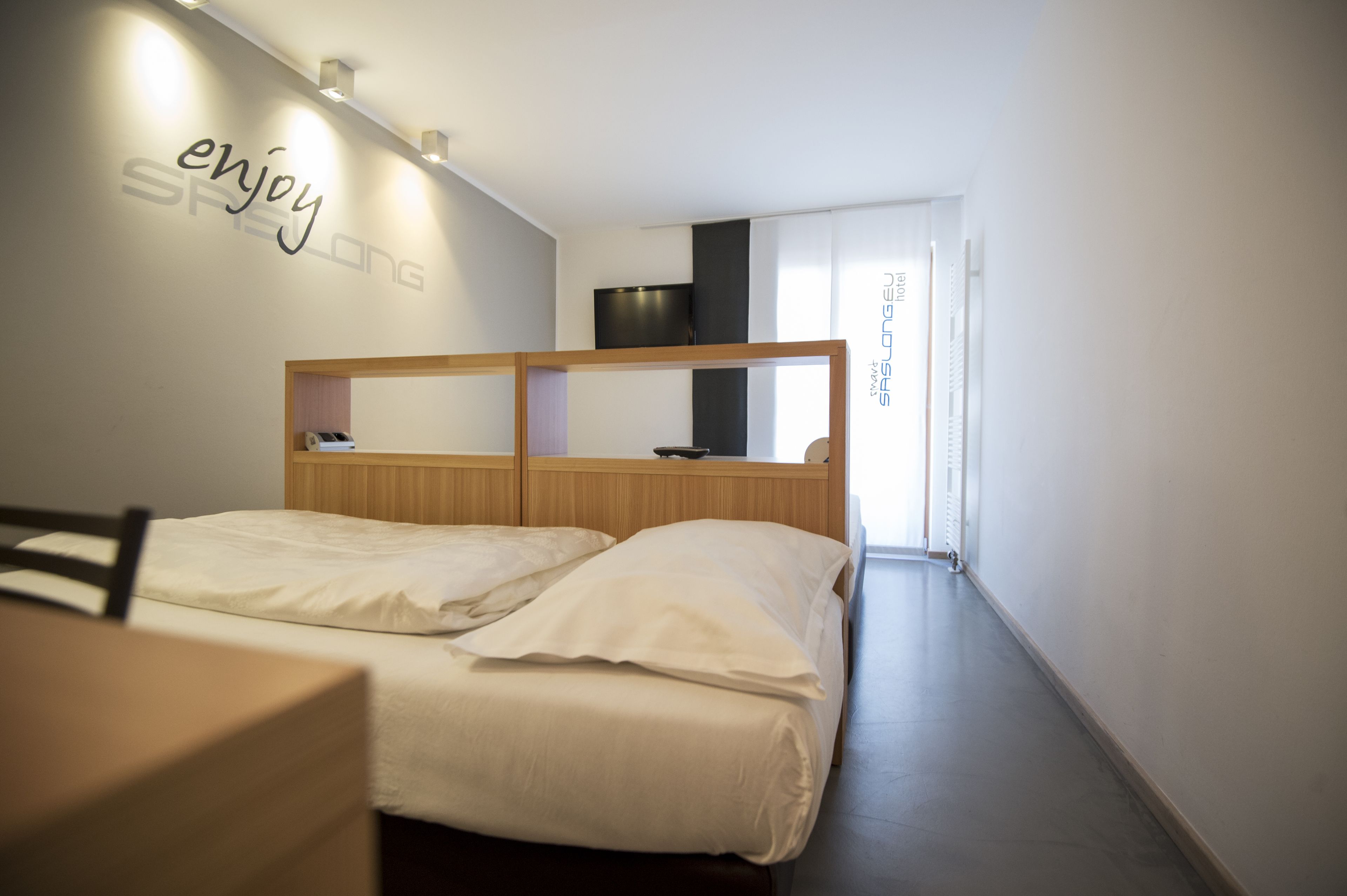 Una habitación de hotel moderna con cabecera de madera, cama con sábanas blancas, una mesita pequeña y un televisor montado en la pared.