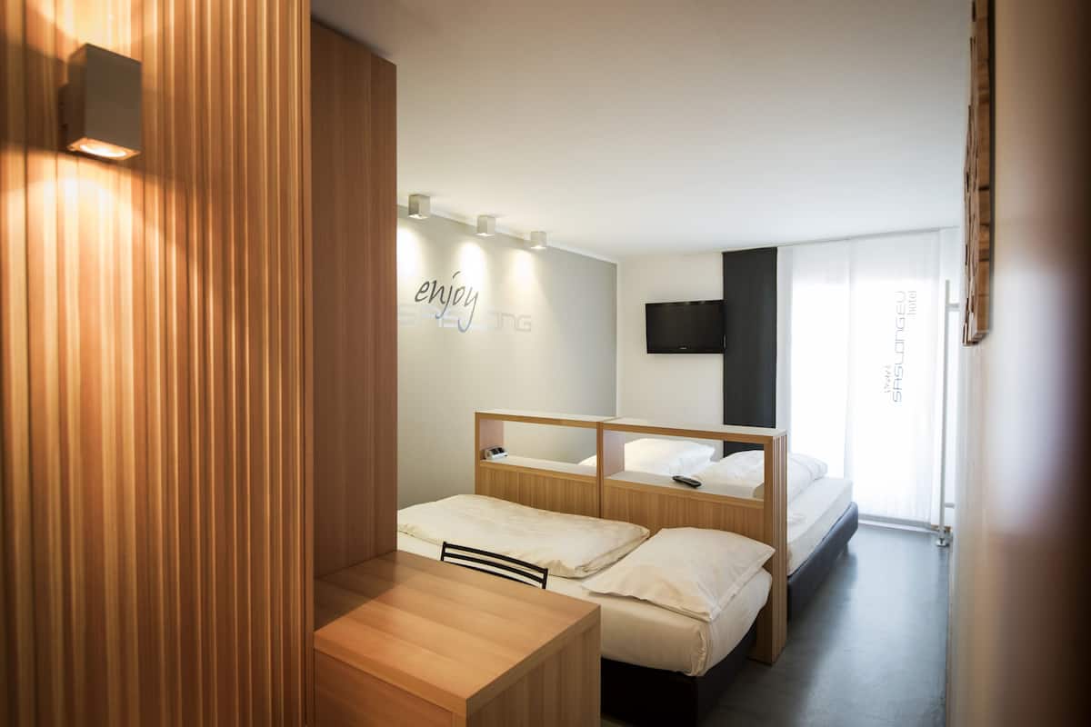 A modern hotel room with two beds, a wooden desk, a mounted TV, and a wall-mounted light fixture.