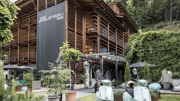 Un hôtel à la façade en bois, sur laquelle on peut lire l’inscription « Hôtel SHLONGEU ». Deux scooters anciens sont garés devant.