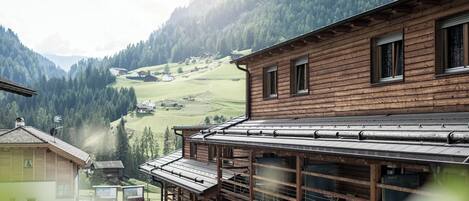 A wooden building with a sloped roof, large windows, and a view of a green valley and mountains in the background.