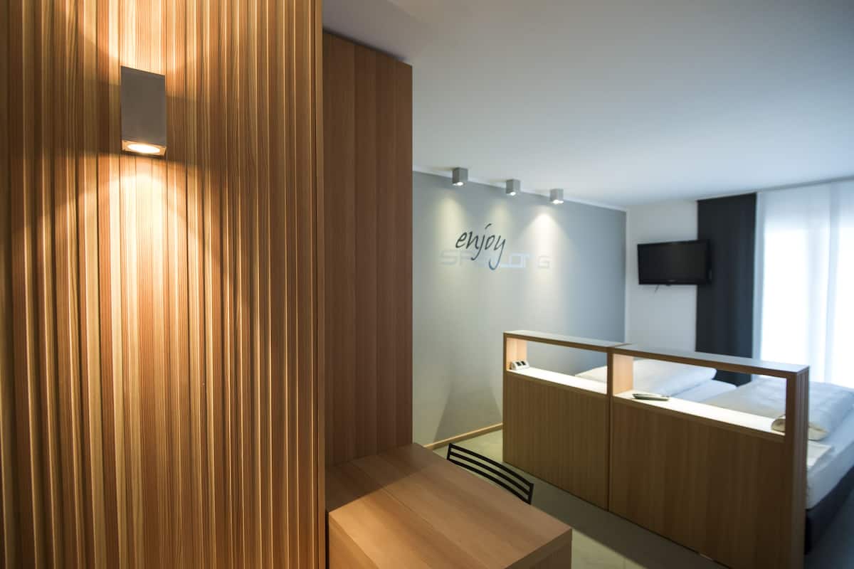 A modern hotel room with a wooden accent wall, a bed with a headboard, a bedside table, and a wall-mounted TV.