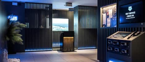 A modern hotel lobby with a reception desk, a self-service check-in kiosk, and a large screen displaying a welcome message.