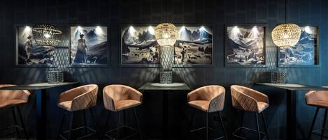 A modern bar area with two tables, each with a unique geometric light fixture and a framed artwork depicting a mountainous landscape.