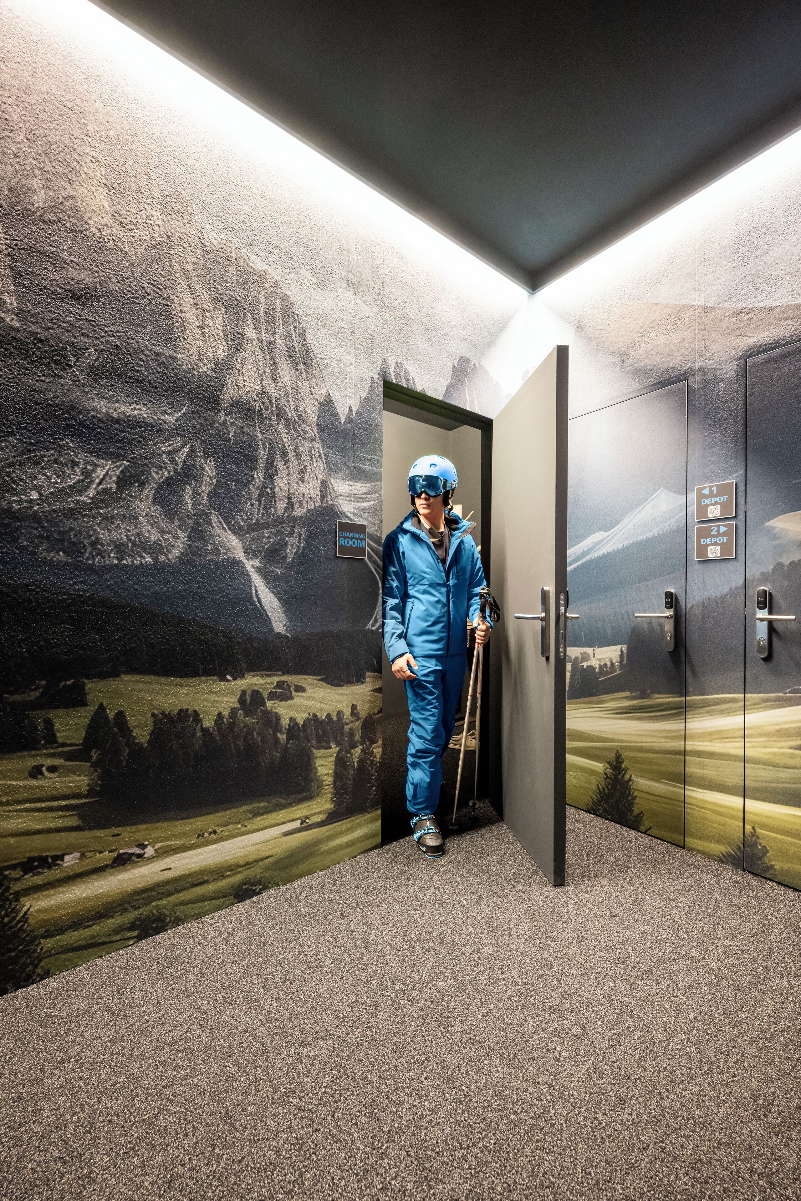Une personne vêtue d’une combinaison de protection bleue se tient dans une pièce ornée d’une fresque sur le thème du ski. On y voit une porte portant l’inscription « Vestiaire » et un panneau indiquant « 2e dépôt ».