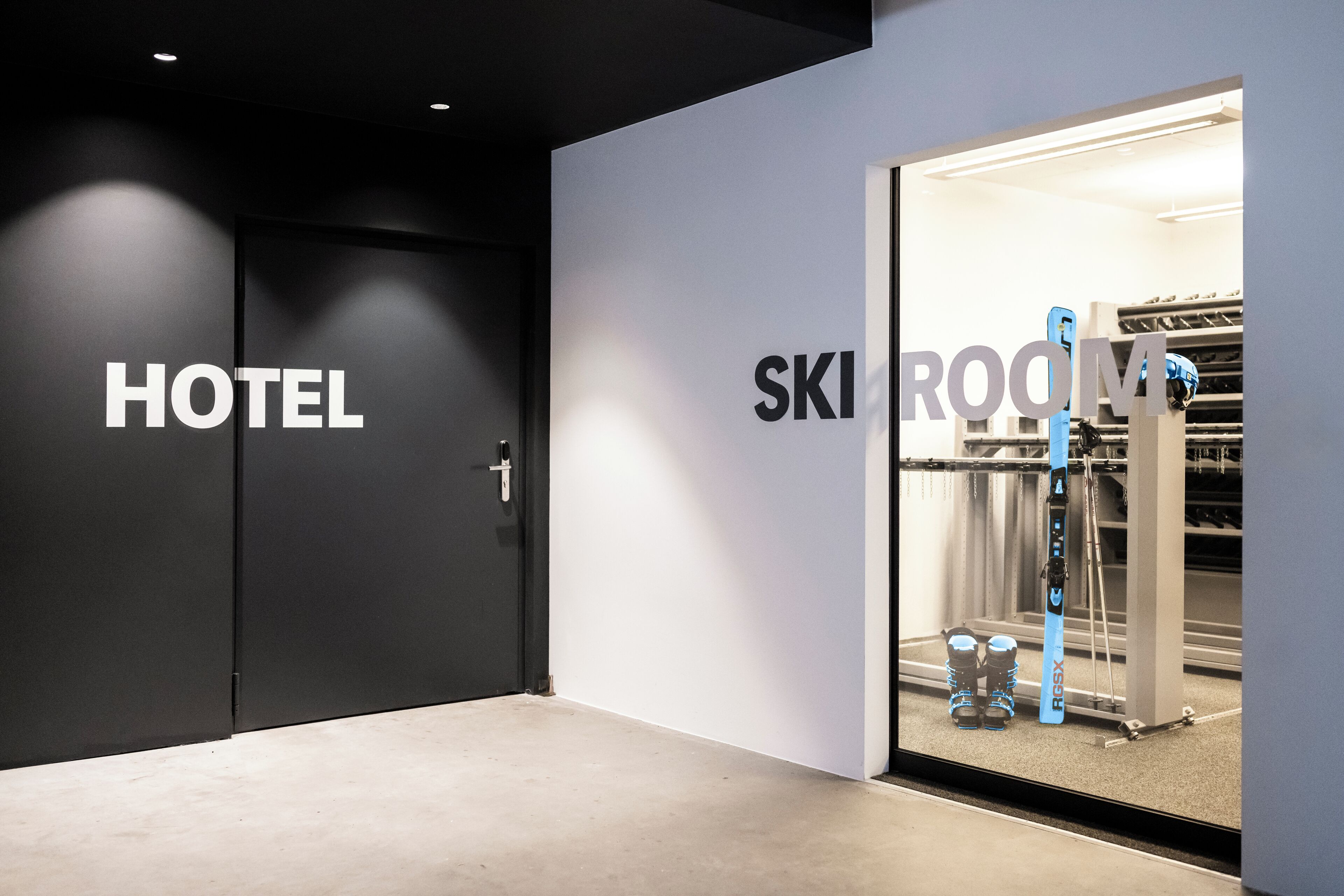A hotel entrance with a sign reading "HOTEL" and a glass door leading to a ski room with skis and a ski lift visible.
