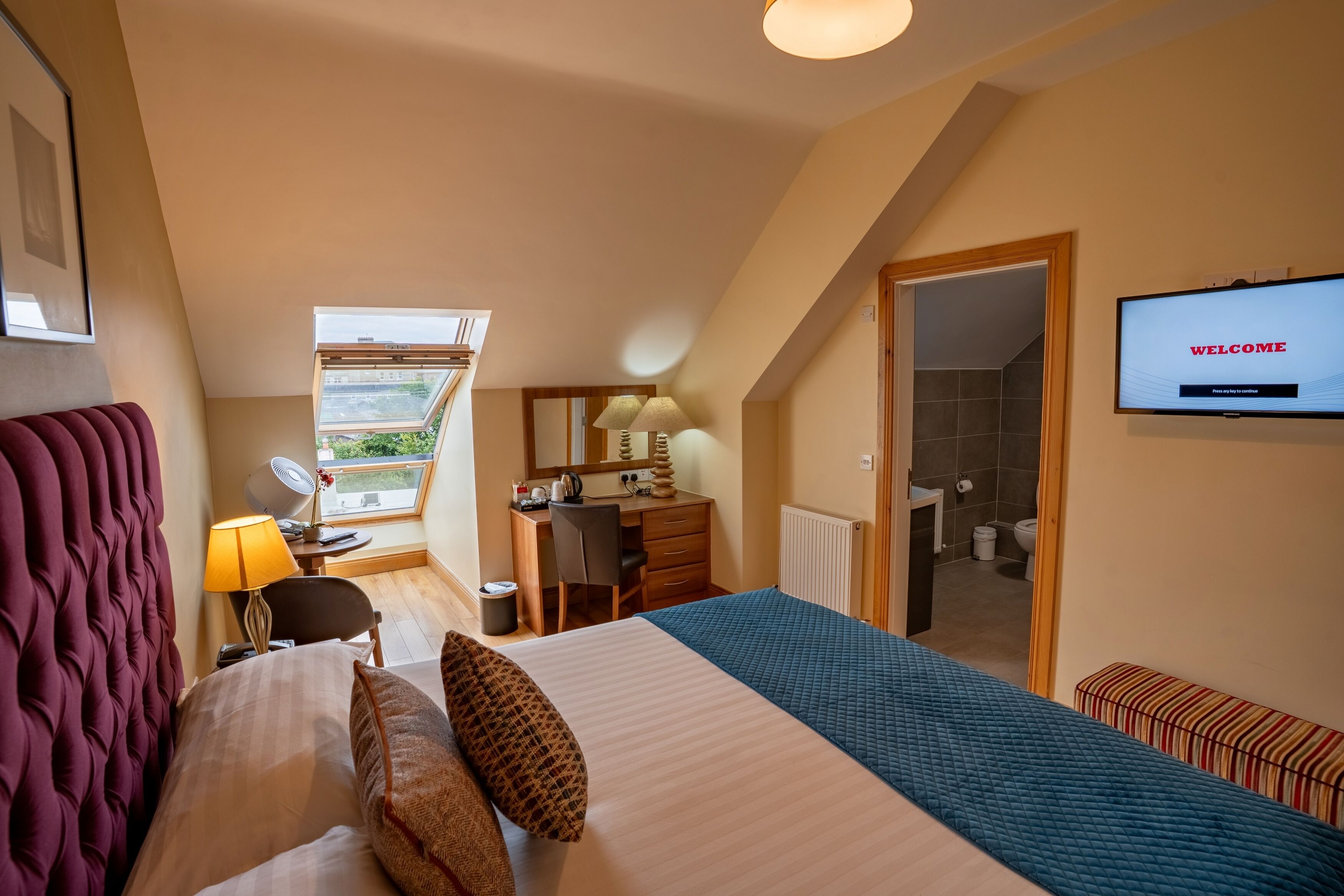 Un dormitorio con cama, escritorio, silla, lámpara, televisión y ventana con vistas.